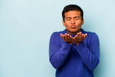 Genç İspanyol adam mavi arka planda izole edilmiş dudaklar kıvrılıyor ve hava öpücüğü göndermek için avuç içleri tutuyor..