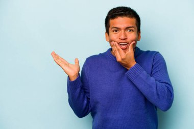 Mavi arka planda izole edilmiş genç İspanyol bir adam avucunun içinde fotokopi alanı tutuyor, el üstünde tutuyor. Şaşırmış ve mutlu..