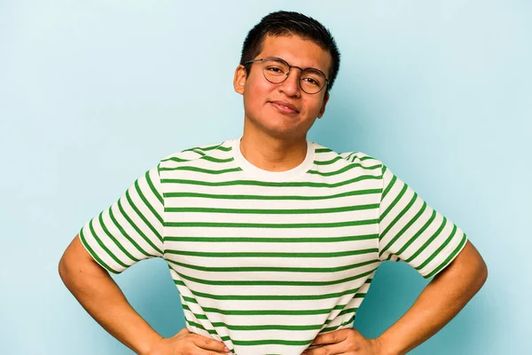 Latin kökenli genç adam mavi arka planda izole edilmiş. Kendine güveniyor. Kalçaları tutuyor..