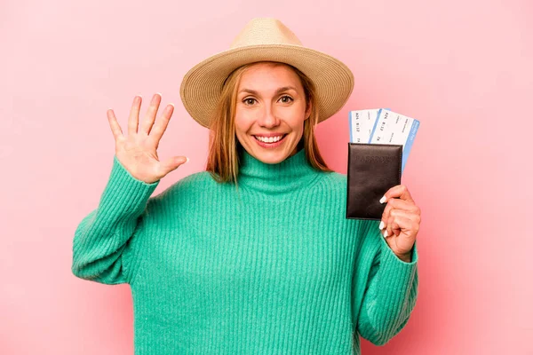 Pembe arka planda izole edilmiş pasaport taşıyan beyaz bir kadın gülümseyerek beş numarayı parmaklarıyla gösteriyor..