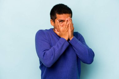 Genç İspanyol adam mavi arka planda izole edilmiş. Korkmuş ve gergin parmaklarıyla göz kırp..