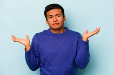 Latin kökenli genç bir adam, mavi arka planda izole edilmiş şüphe içinde ve sorgulama sırasında omuz silkiyor..