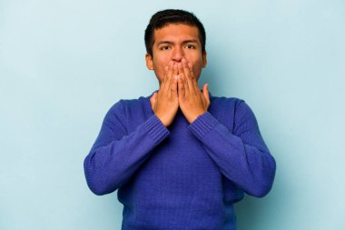 Mavi arka planda izole edilmiş genç İspanyol adam elleriyle ağzını kapatırken şok oldu..