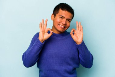 Genç İspanyol adam mavi arka planda izole edilmiş neşeli ve kendine güvenen iyi bir hareket sergiliyor..