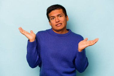 Genç İspanyol bir adam mavi arka planda izole edilmiş. Kafası karışmış ve omuz silkiyor. Kopya alanını tutmak için..