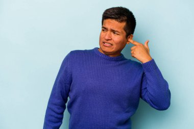 Mavi arka planda izole edilmiş genç İspanyol adam kulaklarını elleriyle kapatıyor..