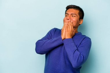 Genç İspanyol bir adam mavi arka planda izole edilmiş bir şekilde bir şeye gülüyor, elleriyle ağzını kapatıyor..