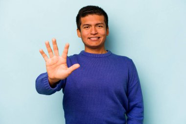 Genç İspanyol adam mavi arka planda izole edilmiş gülümsüyor beş numarayı parmaklarıyla gösteriyor..