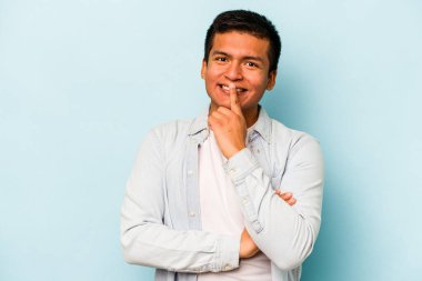 Genç İspanyol adam mavi arka planda izole edilmiş mutlu ve kendinden emin gülümsüyor, elleriyle çenesine dokunuyor..