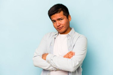 Latin asıllı genç adam mavi arka planda izole edilmiş. Mutsuz bir şekilde kameraya bakıyor. İğneleyici bir ifadeyle..