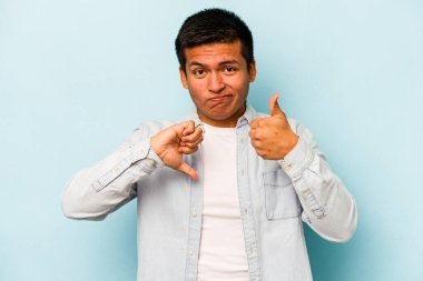 Latin kökenli genç adam mavi arka planda izole edilmiş, baş parmağını kaldırıyor, zor bir seçim konsepti.