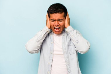 Genç İspanyol bir adam mavi arka planda izole edilmiş kulaklarını elleriyle kapatıyor çok fazla ses duymamaya çalışıyor..