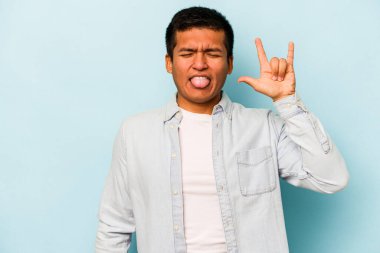 Mavi arka planda izole edilmiş genç İspanyol adam parmaklarıyla kaya hareketi gösteriyor.