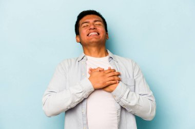 Mavi arka planda izole olmuş genç İspanyol adam gülüyor, ellerini yüreğine koyuyor, mutluluk kavramını koruyor..