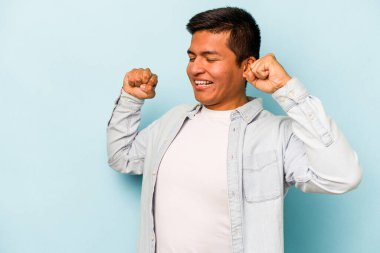 Genç İspanyol adam mavi arka planda izole edilmiş özel bir günü kutluyor, atlıyor ve enerjiyle kollarını kaldırıyor..