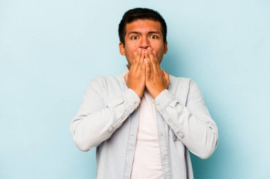 Genç İspanyol bir adam mavi arka planda izole edilmiş, ağzı kapalı ve elleri endişeli görünüyor..