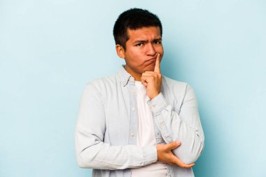 Latin kökenli genç bir adam mavi arka planda izole edilmiş. Bir strateji planlıyor, bir iş yolu düşünüyor..