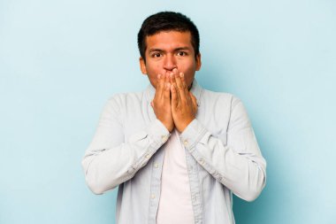 Mavi arka planda izole edilmiş genç İspanyol adam şok olmuş, ağzı ellerle kaplanmış, yeni bir şey keşfetmek için sabırsızlanıyor..