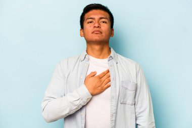 Genç İspanyol bir adam mavi arka planda izole edilmiş yemin ediyor, elini göğsüne koyuyor..