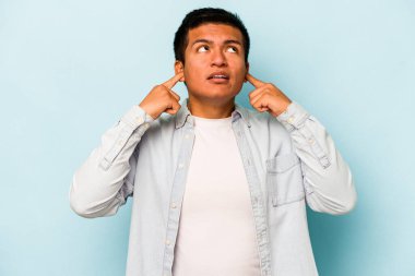 Genç İspanyol bir adam mavi arka planda izole edilmiş parmaklarıyla kulaklarını kapatıyor, gürültülü bir ortam yüzünden stresli ve çaresiz..