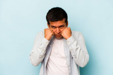 Latin kökenli genç bir adam mavi arka planda izole edilmiş. Yumruk atıyor, sinirleniyor, kavga ediyor..