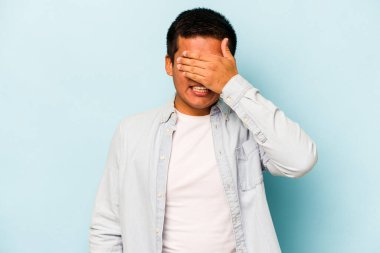 Mavi arka planda izole edilmiş genç İspanyol adam elleriyle gözlerini kapatıyor, geniş bir gülümsemeyle bir sürpriz bekliyor..