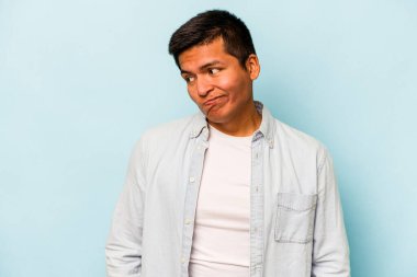 Mavi arka planda izole edilmiş genç bir İspanyol omuz silker ve gözleri açık kalır..