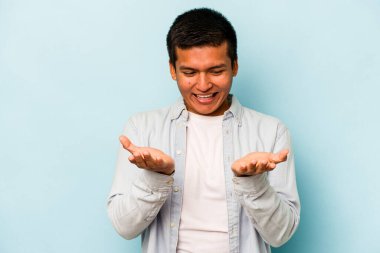 Genç İspanyol bir adam mavi arka planda izole edilmiş. Elinde avuç içi olan bir şeyle kameraya sunuyor..