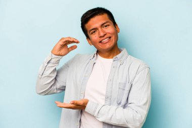 Mavi arka planda izole edilmiş genç İspanyol adam işaret parmaklarıyla küçük bir şey tutuyor, gülümsüyor ve kendinden emin..