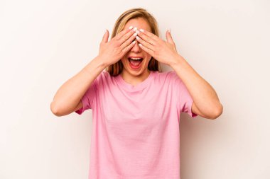 Beyaz arka planda soyutlanmış beyaz tenli genç bir kadın elleriyle gözlerini kapatıyor, geniş bir gülümsemeyle bir sürpriz bekliyor..