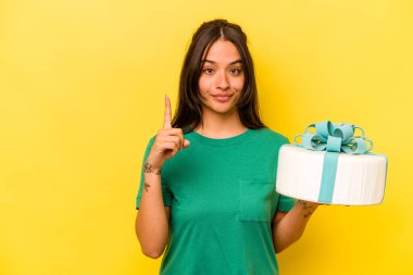 Sarı arka planda izole edilmiş doğum günü pastası tutan genç İspanyol kadın bir numarayı parmağıyla gösteriyor..