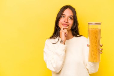Sarı arka planda izole edilmiş spagetti kavanozunu tutan genç İspanyol kadın şüpheli ve şüpheci bir ifadeyle yan yana bakıyor..