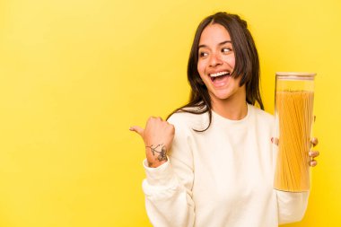 Elinde spagetti kavanozu tutan genç İspanyol kadın sarı arka planda izole edilmiş, baş parmak uzakta, gülen ve kaygısız..
