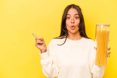 Sarı arka planda izole edilmiş spagetti kavanozunu tutan genç İspanyol kadın yan tarafı işaret ediyor.