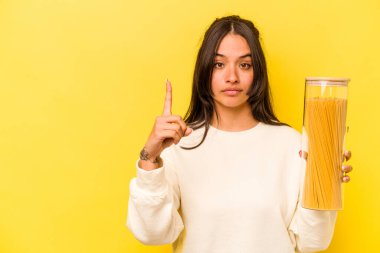 Sarı arka planda izole edilmiş spagetti kavanozu tutan genç İspanyol kadın parmak ile bir numarayı gösteriyor..