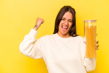 Sarı arka planda izole edilmiş spagetti kavanozunu tutan genç İspanyol kadın zaferden sonra yumruğunu kaldırıyor, kazanan konsept..