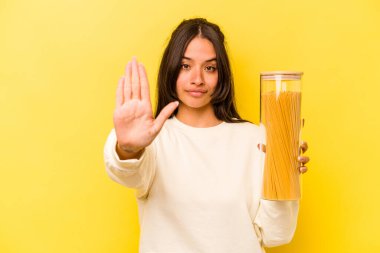 Sarı arka planda izole edilmiş spagetti kavanozunu tutan genç İspanyol kadın elini uzatmış, dur işaretini göstererek seni engelliyor..