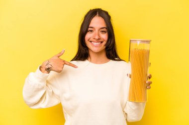 Sarı arka plandaki izole edilmiş spagetti kavanozunu tutan genç İspanyol kadın gömlek fotokopi alanını işaret ediyor, gururlu ve kendinden emin.