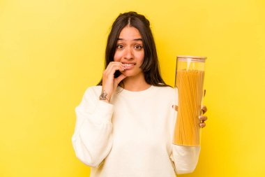 Sarı arka planda izole edilmiş, tırnak yiyen, gergin ve endişeli bir spagetti kavanozu tutan genç İspanyol kadın..