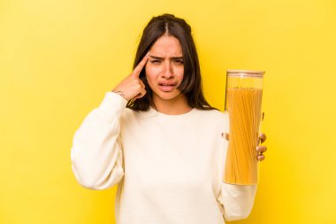 Sarı arka planda izole edilmiş spagetti kavanozunu tutan genç İspanyol kadın işaret parmağıyla yaptığı hayal kırıklığı jestini gösteriyor..