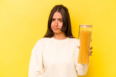 Sarı arka planda izole edilmiş spagetti kavanozunu tutan genç İspanyol kadın şaşkın ve kararsız hissediyor..