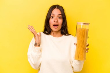 Sarı arka planda izole edilmiş spagetti kavanozunu tutan genç İspanyol kadın şaşırdı ve şok oldu..