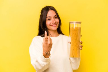 Sarı arka planda izole edilmiş spagetti kavanozunu tutan genç İspanyol kadın sanki seni davet ediyormuş gibi seni işaret ediyor..