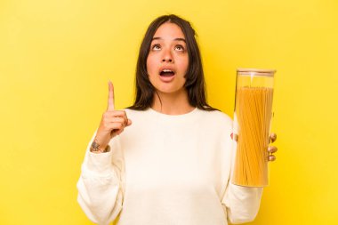 Sarı arka planda izole edilmiş spagetti kavanozu tutan genç İspanyol kadın ağzı açık bir şekilde üst tarafı işaret ediyor..