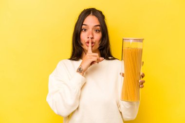 Sarı arka planda izole edilmiş spagetti kavanozunu tutan genç İspanyol kadın ya sır saklıyor ya da sessizlik istiyor..