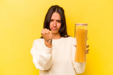 Sarı arka planda izole edilmiş spagetti kavanozu tutan genç İspanyol kadın kameraya yumruk gösteriyor, agresif yüz ifadesi..