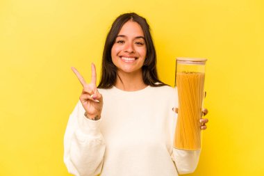 Sarı arka planda izole edilmiş spagetti kavanozu tutan genç İspanyol kadın iki numarayı parmaklarıyla gösteriyor..