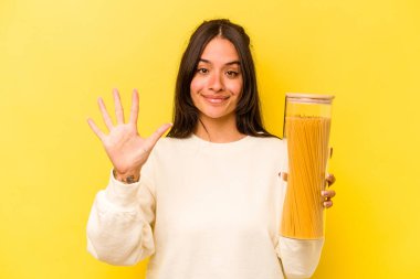 Sarı arka planda izole edilmiş spagetti kavanozunu tutan genç İspanyol kadın gülümseyerek beş numarayı parmaklarıyla gösteriyor..