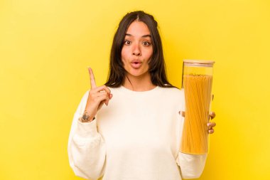 Sarı arka planda izole edilmiş spagetti kavanozunu tutan genç İspanyol kadının harika bir fikri var, yaratıcılık kavramı..