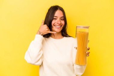 Sarı arka planda izole edilmiş spagetti kavanozu tutan genç İspanyol kadın cep telefonu hareketini parmaklarıyla gösteriyor..